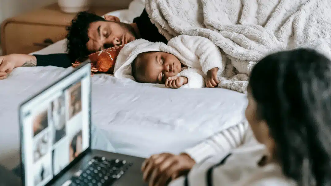 A foster care family relaxing together in a cozy bedroom; an adult parent on bed cuddles a child in white, while another parent works on a laptop nearby.