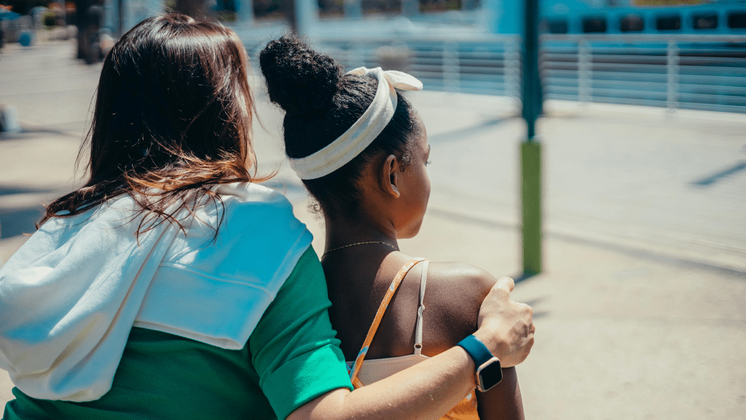 Adult and child are seen from behind, with adult person placing an arm around the child's shoulders as they stand outdoors near a railing on a sunny day.
