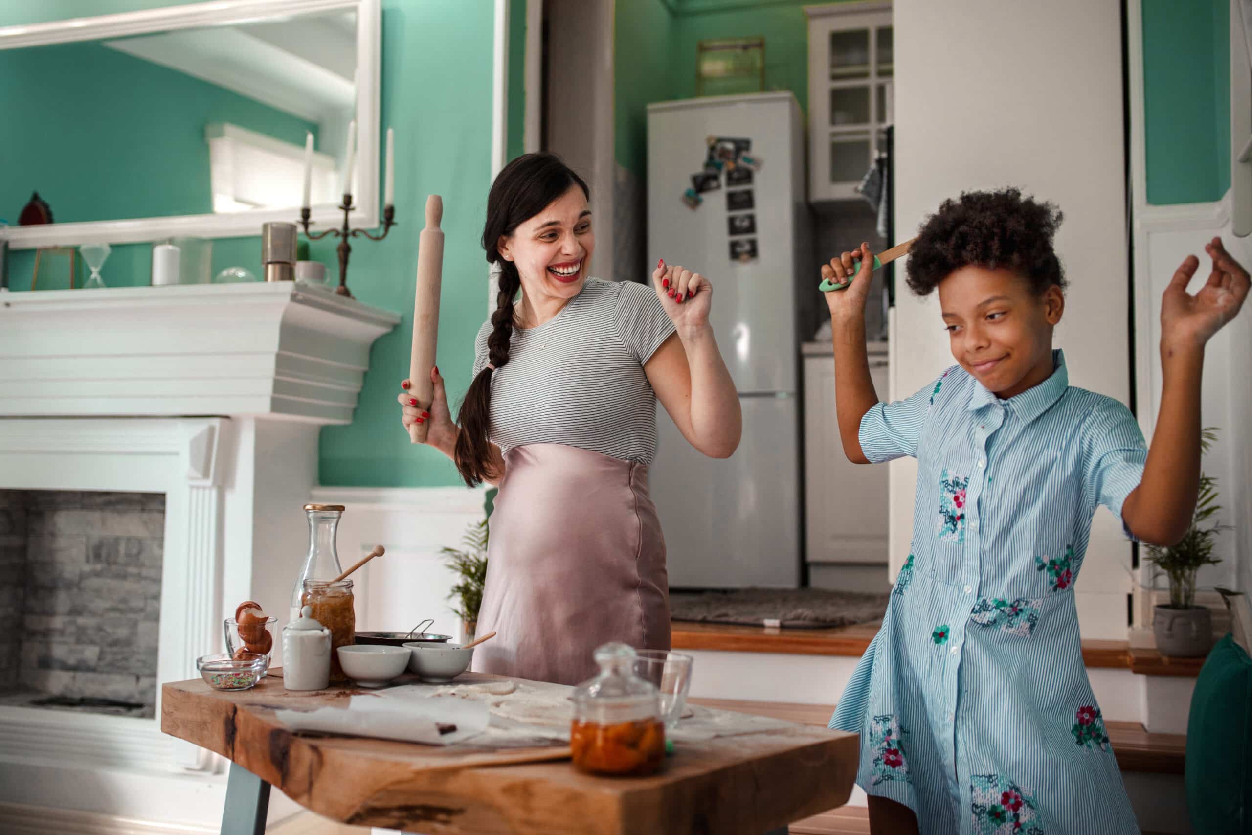 A joyful woman and child dance in a bright kitchen, playfully holding utensils. The table is set with cooking items, and the atmosphere is lively and cheerful.