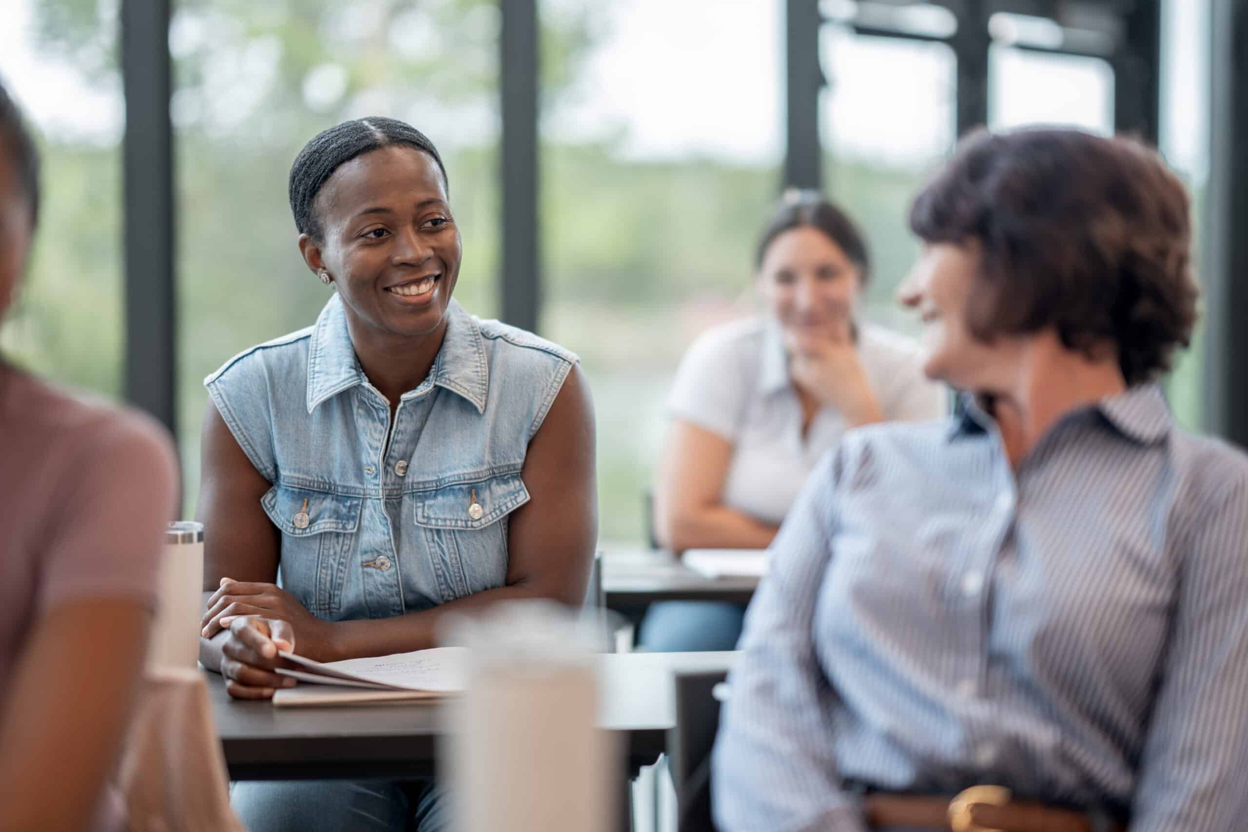 A smiling woman in a denim vest engages with a classmate in a bright, airy classroom. Others in the background are seated, fostering a friendly, relaxed atmosphere.
