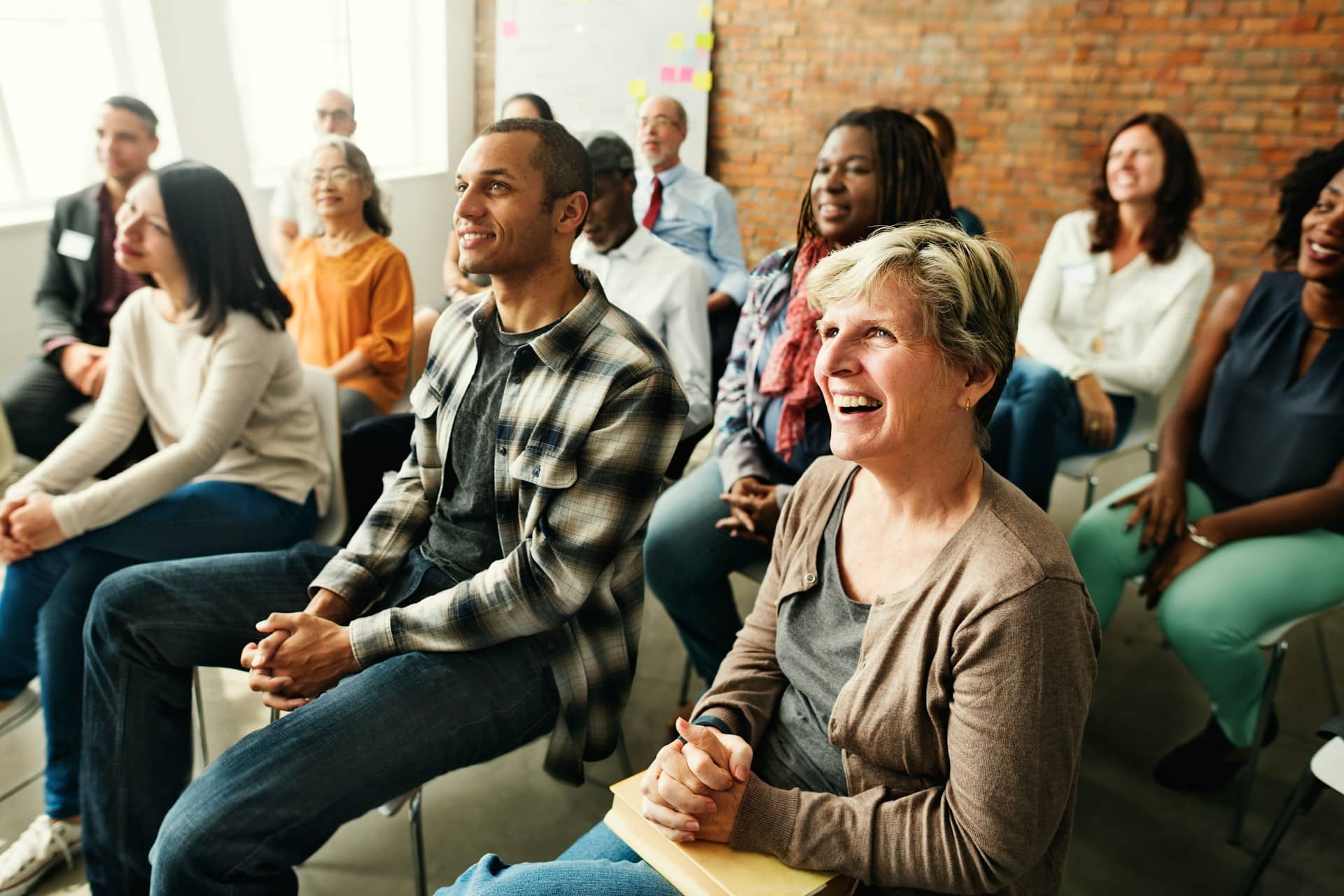 A diverse group of adults sits in a bright room with brick walls, listening attentively and smiling during a relaxed, welcoming group session.