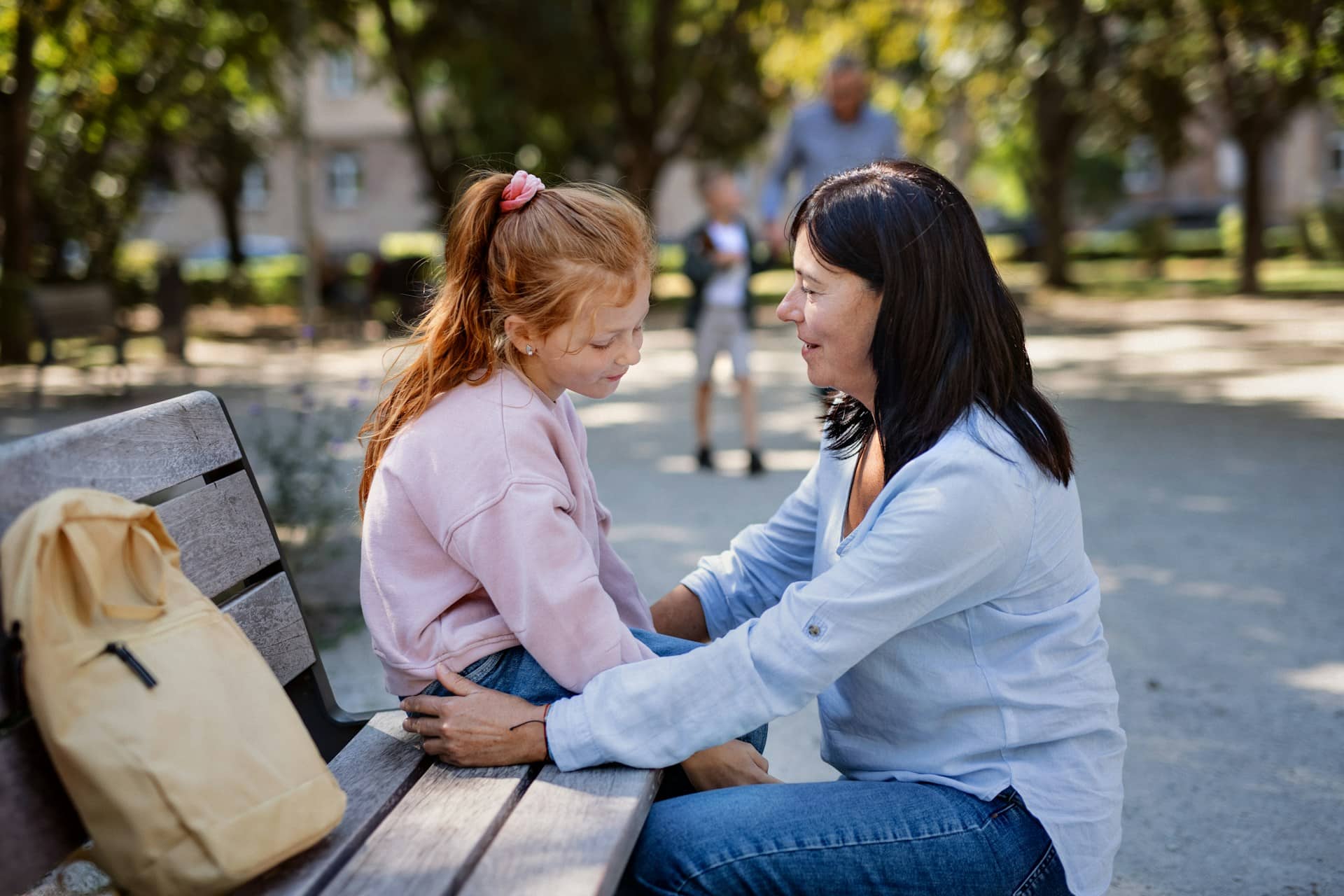 A woman leans toward a young girl on a park bench, holding her hands in a gentle, reassuring moment; a backpack and blurred parkgoers fill the background.