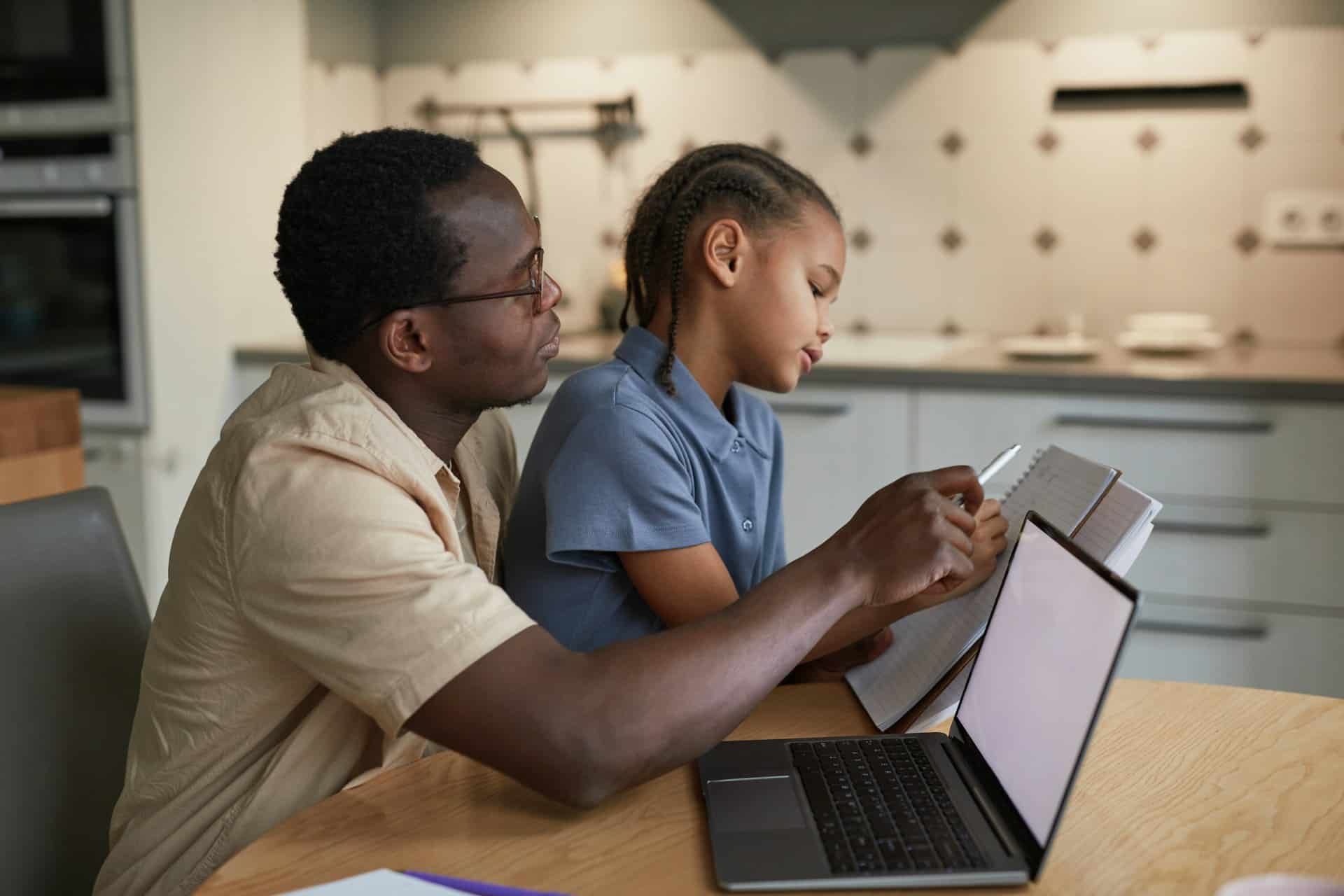 An adult man guides a young girl writing in a notebook beside an open laptop at a kitchen table, sharing a calm, focused study moment.