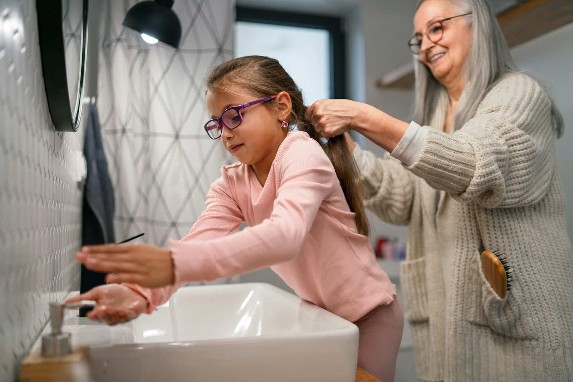 A smiling older woman gathers a young girl's hair as the girl reaches for a bathroom faucet over a white sink, creating a warm, caring home scene.