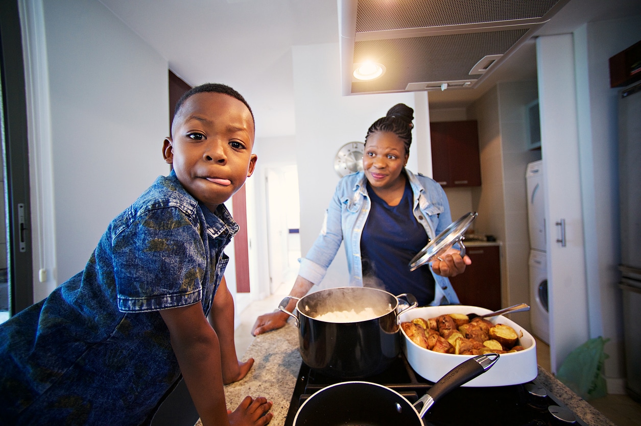 A boy leans toward the camera in a kitchen while a woman cooks beside simmering pots and a pan of food, creating a warm family scene.