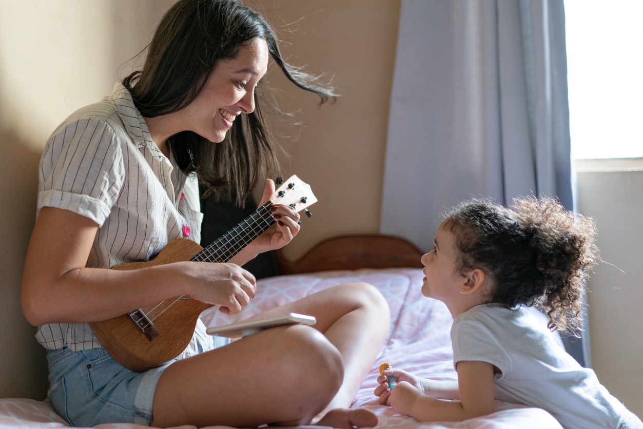 A smiling mother strums a small ukulele on a bed as a young child watches, sharing a cheerful, intimate music moment in a sunlit bedroom.