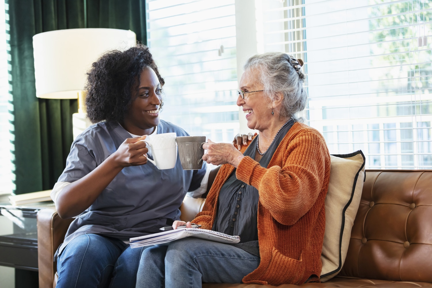 A younger Black woman and an older white woman smile over mugs on a sofa, with a notebook on their laps in a bright, welcoming living room.