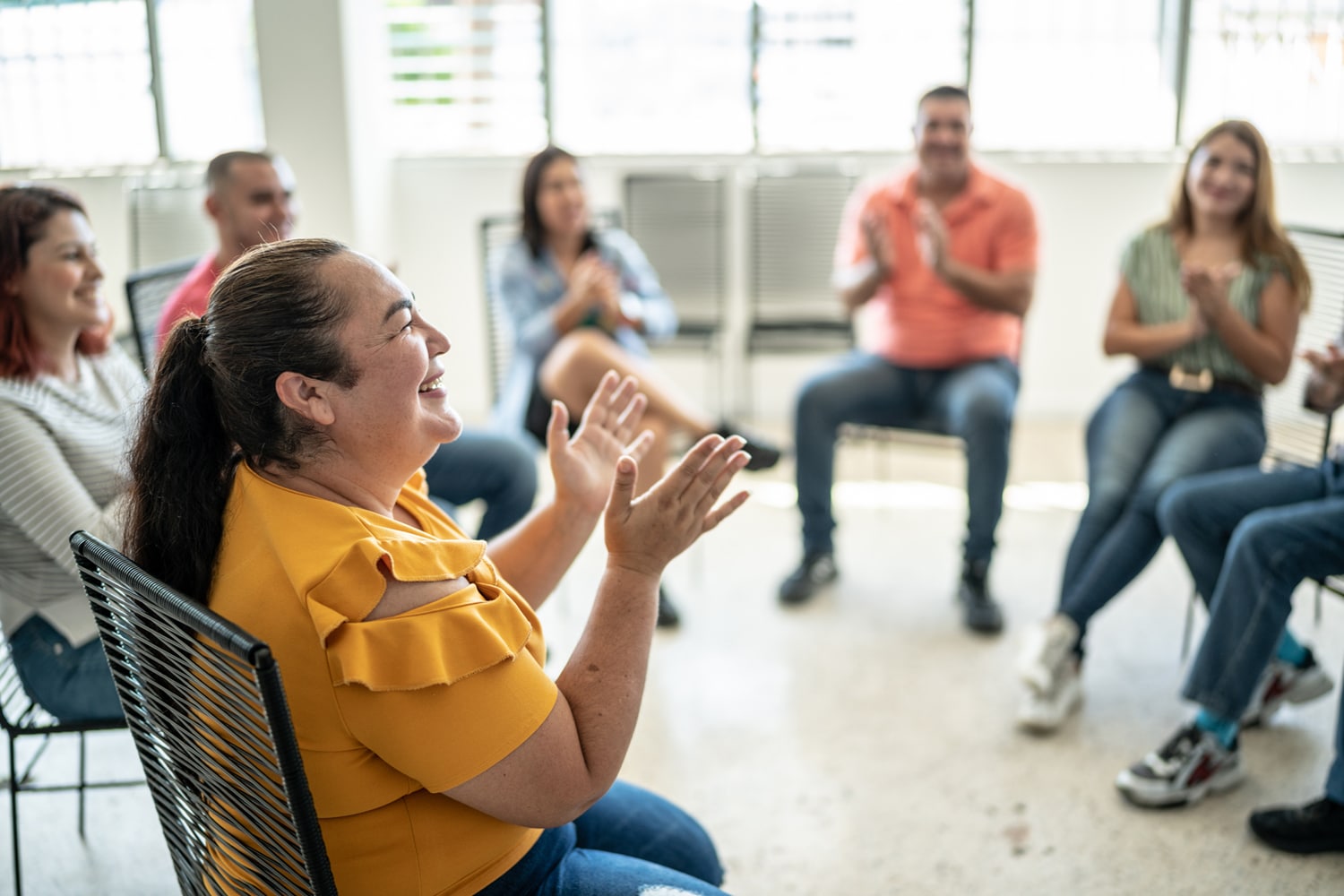 A woman in a mustard-yellow shirt laughs and claps in a circle with smiling adults in a bright room, creating a warm, supportive group atmosphere.