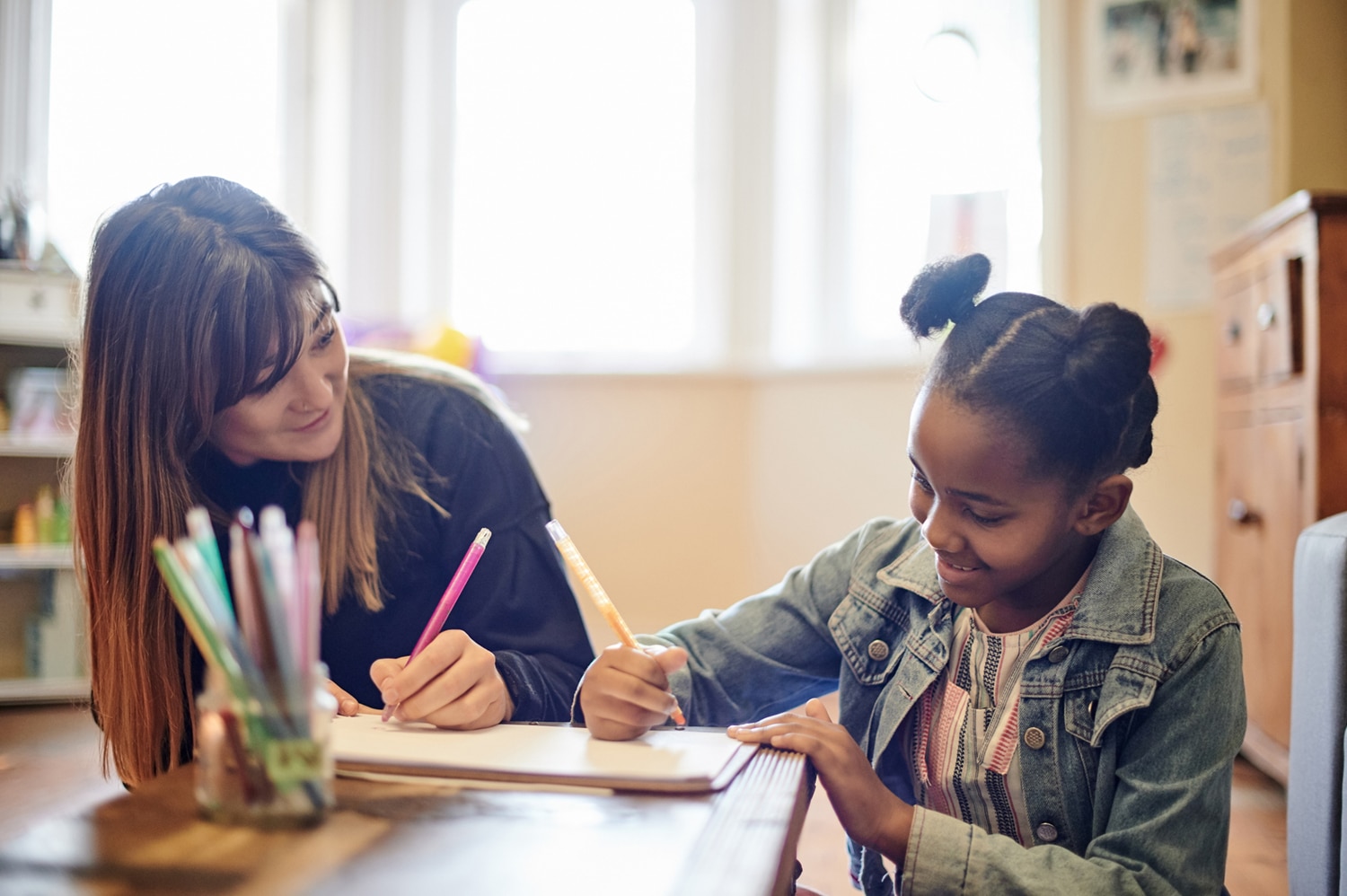 A teacher and young girl write together at a classroom table, surrounded by pencils and notebooks; the child’s smile creates a warm, encouraging mood.