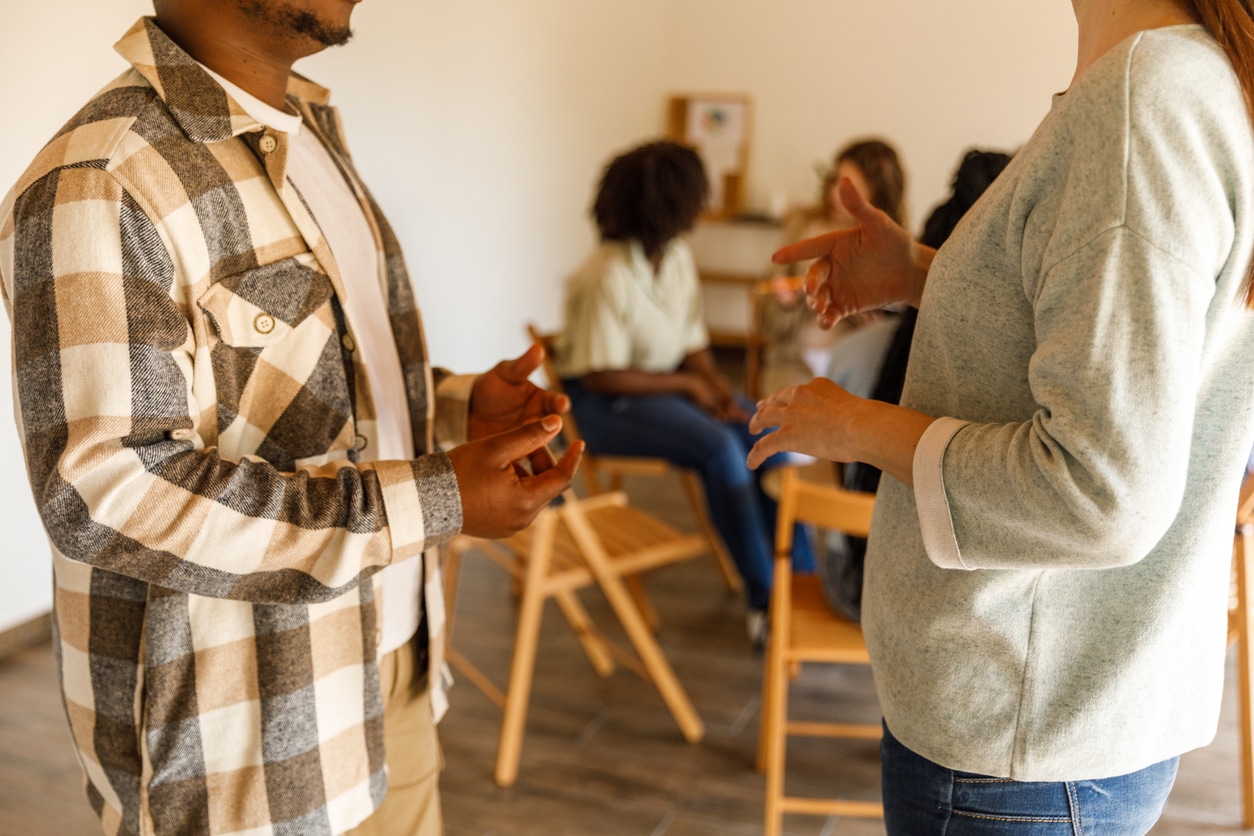 Two people converse with animated hand gestures in a warm, casual room, while others sit on wooden chairs in the background; the scene feels friendly and relaxed.