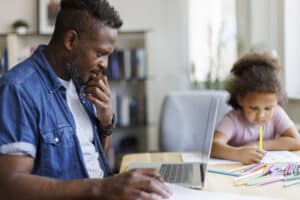 A thoughtful father watches his young daughter write beside an open laptop and colorful pencils in a bright, calm home study space.