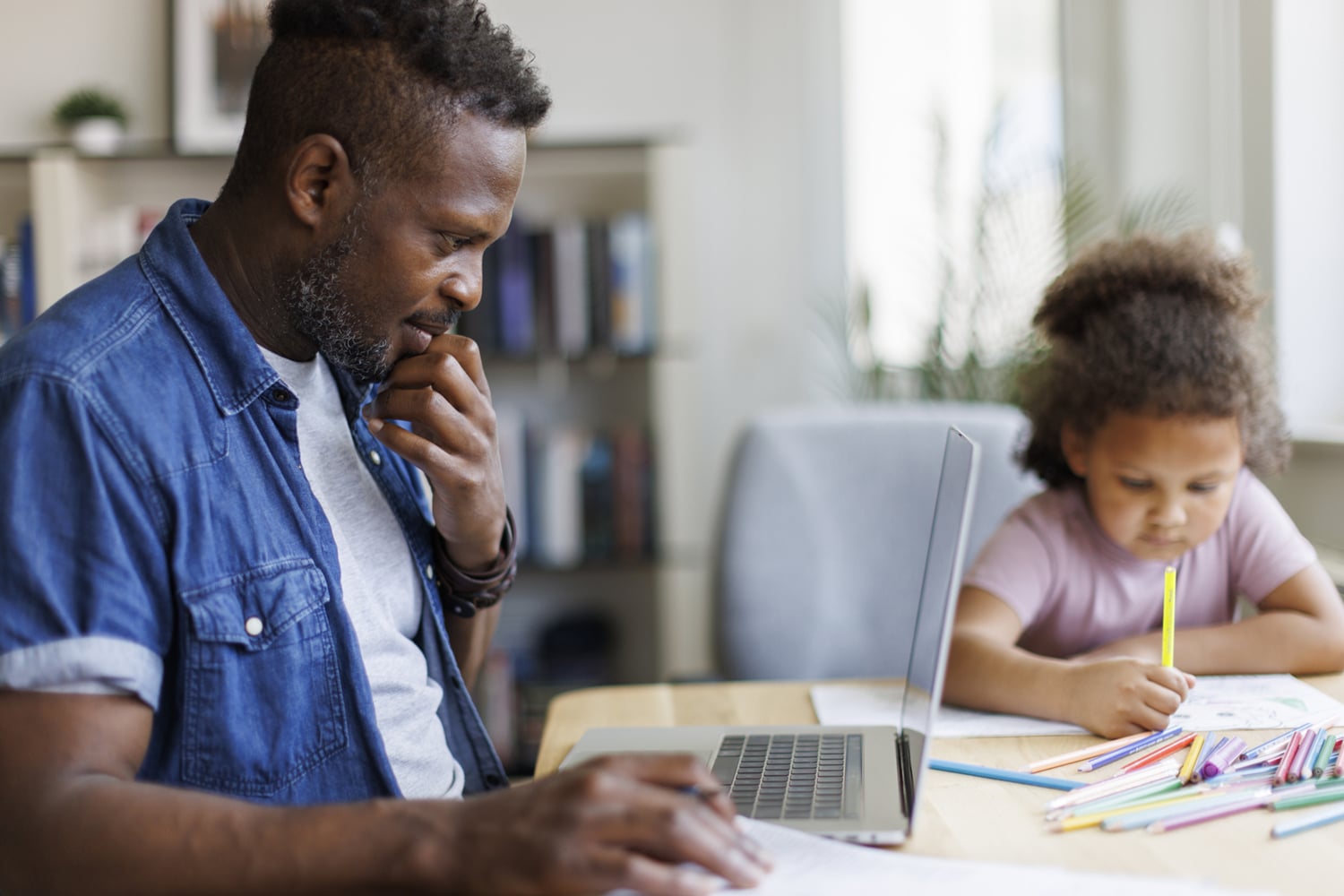 A thoughtful father watches his young daughter write beside an open laptop and colorful pencils in a bright, calm home study space.