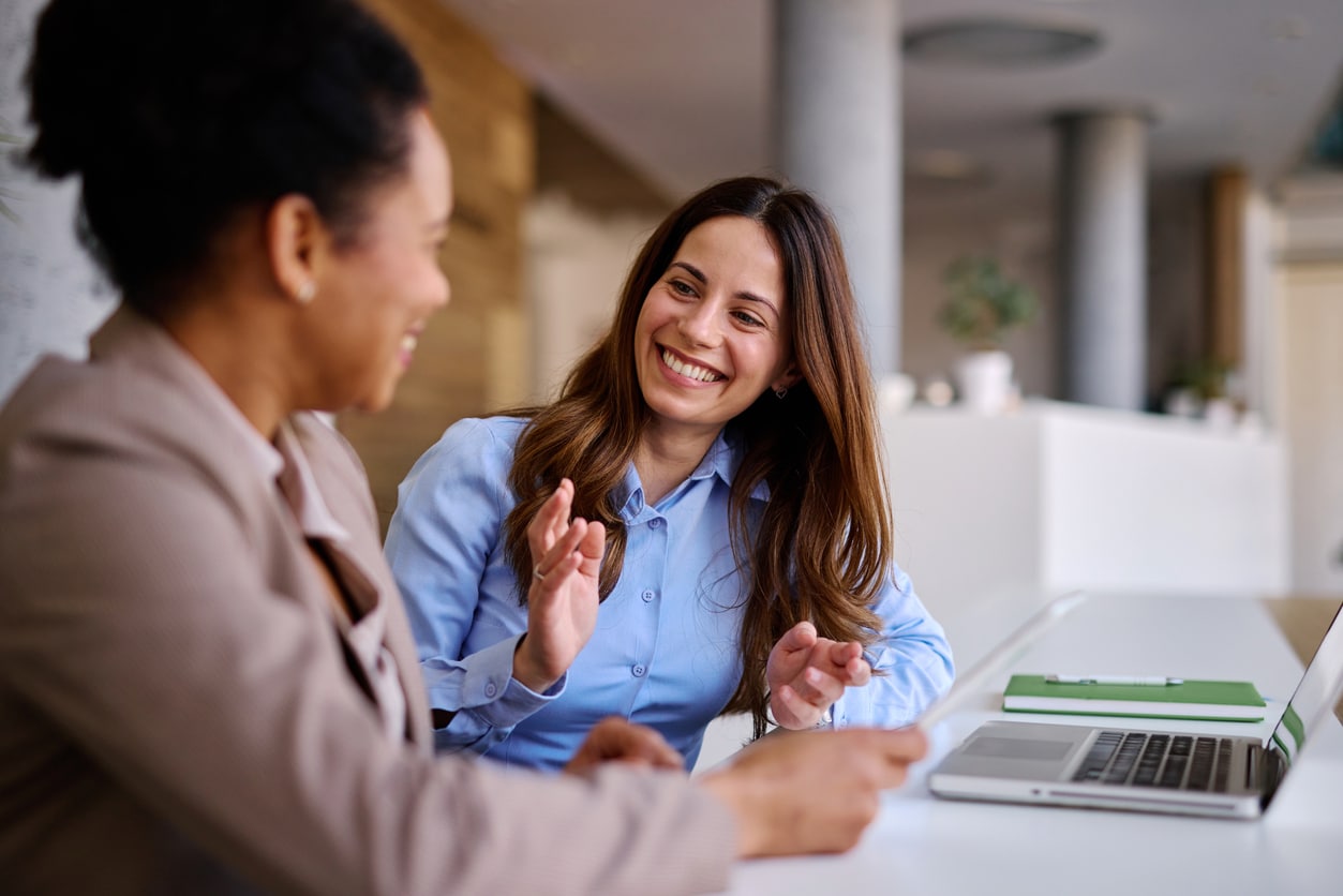 Two colleagues smile and chat across an office desk, one gesturing as an open laptop and papers sit nearby; the bright, relaxed workplace feels friendly and collaborative.